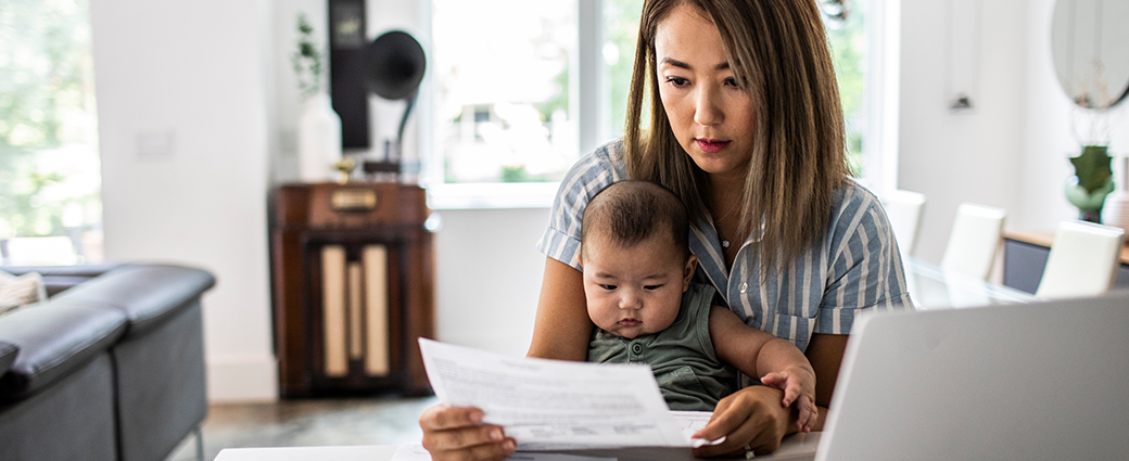 A woman sitting in a living room, holding a baby and looking at a piece of paper.